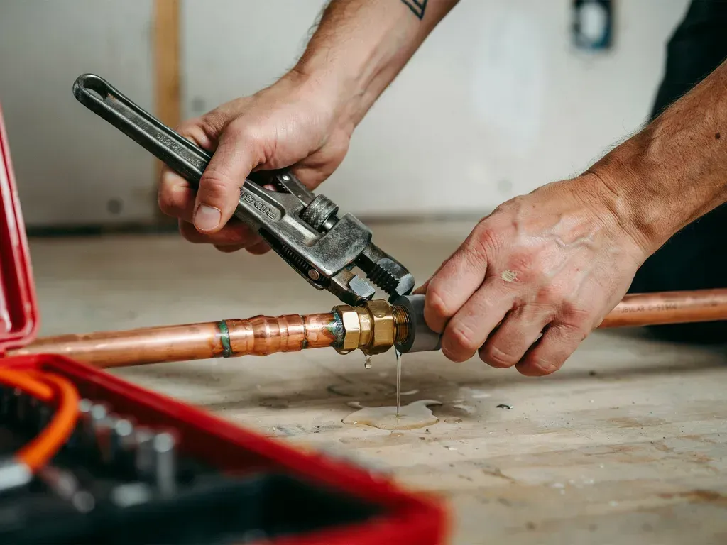 Hands using a pipe wrench to tighten a copper pipe fitting on a floor.