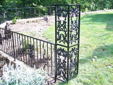 Black wrought-iron fence and corner post with floral design, surrounding a flowerbed and grassy lawn.