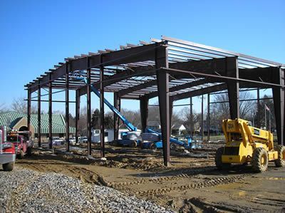 Steel frame of a building under construction, with heavy machinery on a dirt lot, against a blue sky.