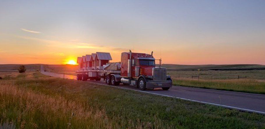 A semi-truck hauling cargo on a rural road at sunset; orange sky, green fields.