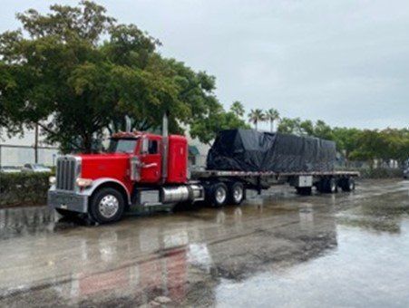 Red semi-truck and trailer hauling a black-covered load on wet pavement, trees in the background.