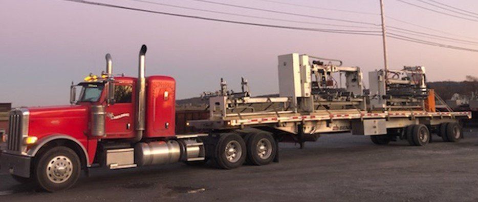 A red semi-truck hauling large machinery on a flatbed trailer in an industrial setting.