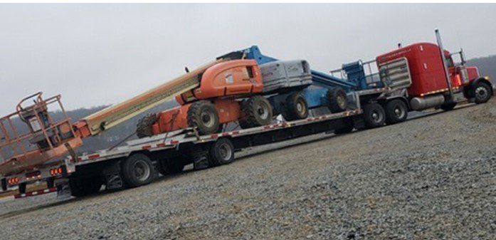 Red semi truck hauling heavy machinery on a flatbed trailer on a gravel road.