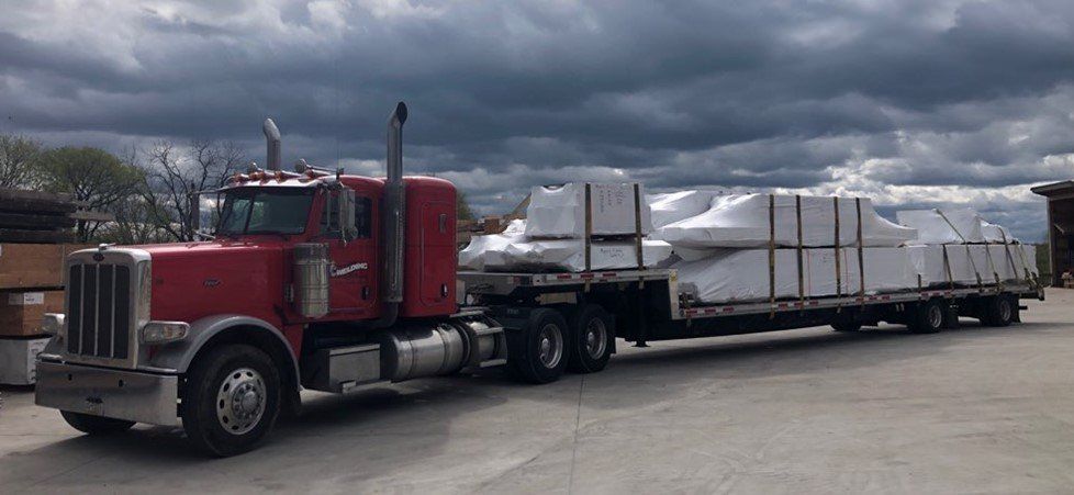 A red semi-truck hauling a flatbed trailer loaded with white, rectangular materials under a cloudy sky.