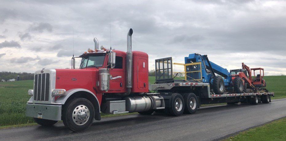 Red semi-truck hauling heavy machinery on a trailer on a rural road under an overcast sky.