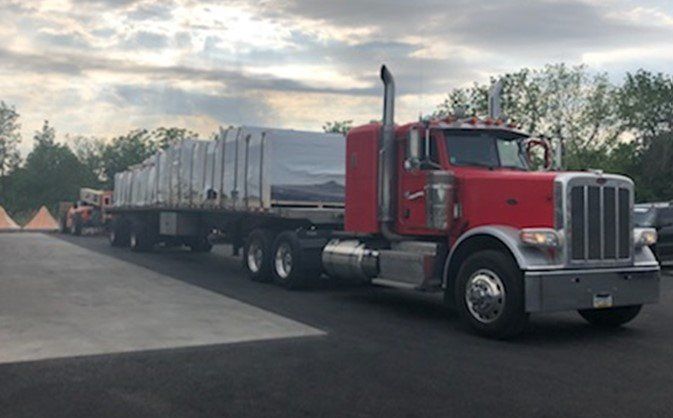 Red semi-truck with flatbed trailer hauling covered cargo in outdoor setting. Cloudy sky in the background.