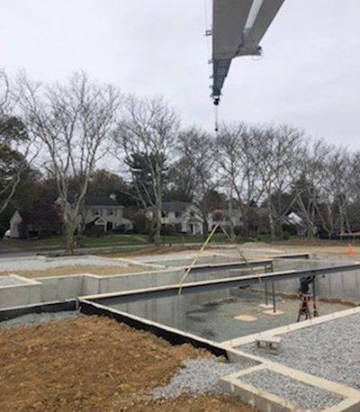 Construction site with concrete foundation, crane overhead, trees, and houses in the background on a cloudy day.