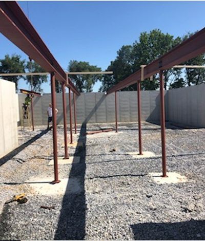 Steel beams supported by vertical posts, construction site. Gravel ground, concrete wall background, blue sky. Man stands near the structure.