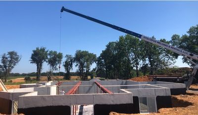 Construction site with crane lifting over a concrete foundation. Blue sky, green trees, and a field in the distance.