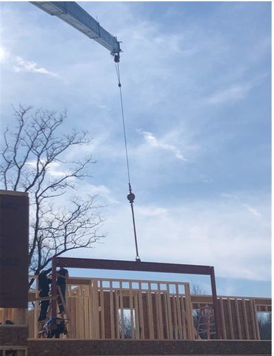 Crane lifting a wooden wall frame during construction on a sunny day.