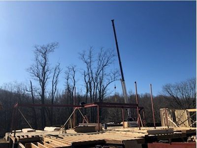 Construction site with crane lifting steel beams; wooden framework in foreground; trees and blue sky.