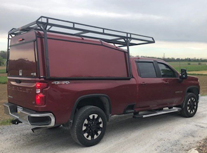 Maroon Chevrolet Silverado truck with a matching camper shell and black roof rack. Parked on a gravel road.