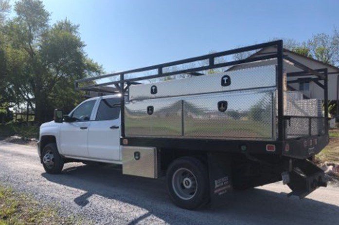 White work truck with a flatbed, toolboxes, and overhead rack parked on a gravel road.