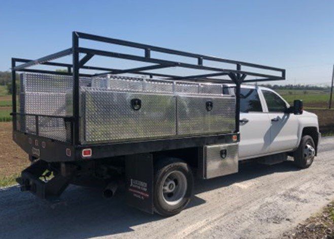 White work truck with black flatbed, toolboxes, and overhead rack on a gravel road.