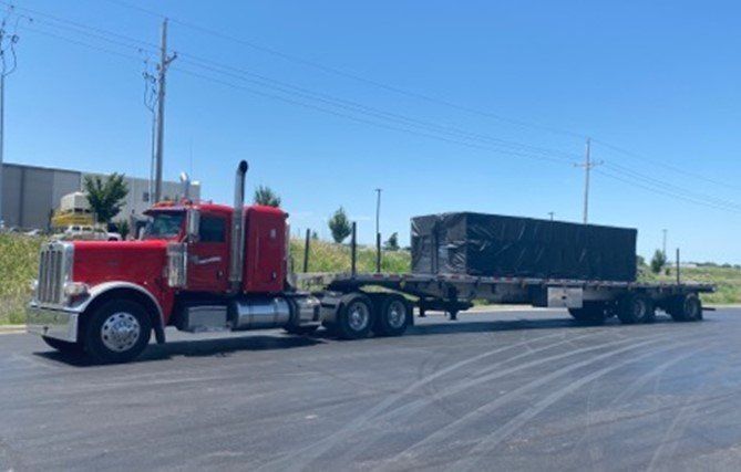 Red semi-truck hauling a black tarp-covered load on a flatbed trailer, parked on asphalt under a blue sky.