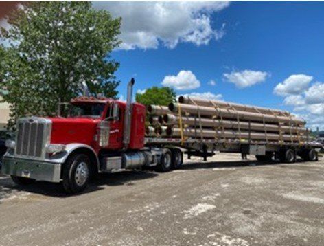 Red semi-truck hauling a trailer loaded with cylindrical pipes on a bright, sunny day.