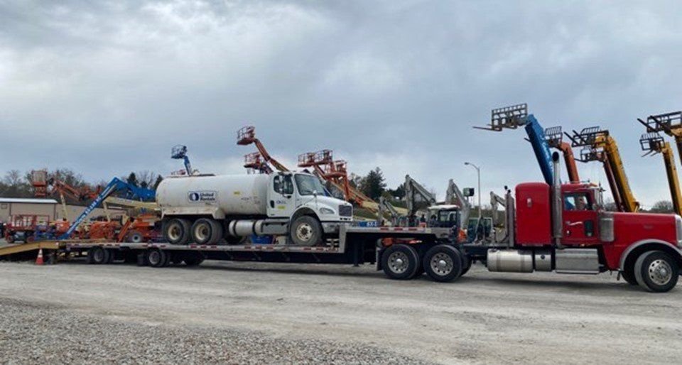 A red semi-truck pulling a flatbed trailer loaded with equipment, including a water tanker and aerial lifts on an overcast day.
