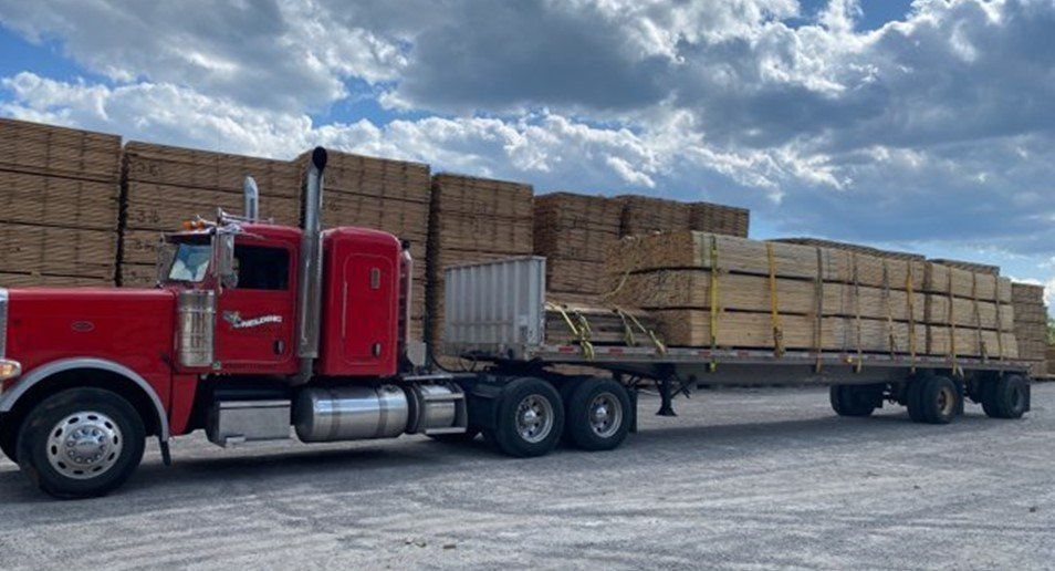 Red semi-truck with flatbed trailer loaded with lumber at a lumberyard on a cloudy day.