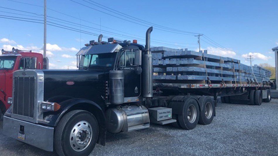 Black semi-truck hauling a flatbed loaded with gray metal plates on a gravel lot under a clear sky.