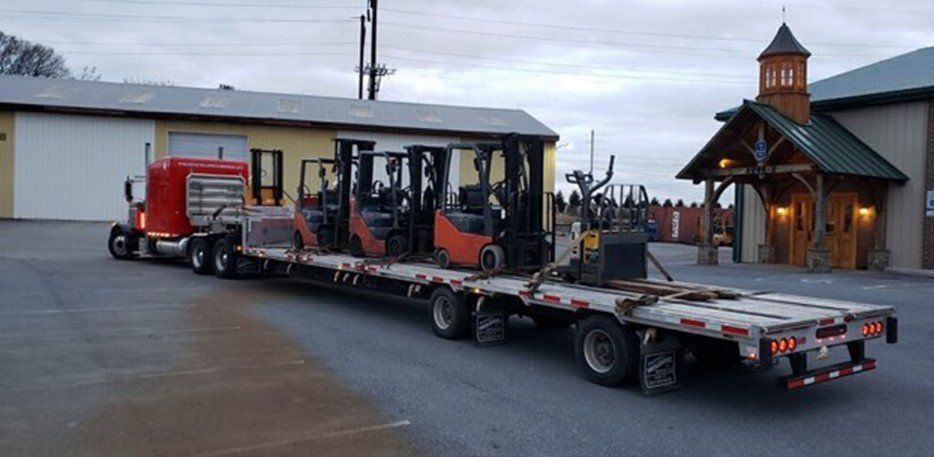 A semi-truck hauling several forklifts on a flatbed trailer in front of a building with a wooden entrance.