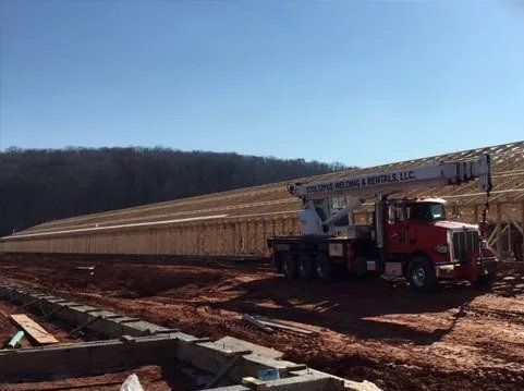 Construction site with a red crane truck lifting materials onto a wooden frame, against a blue sky.