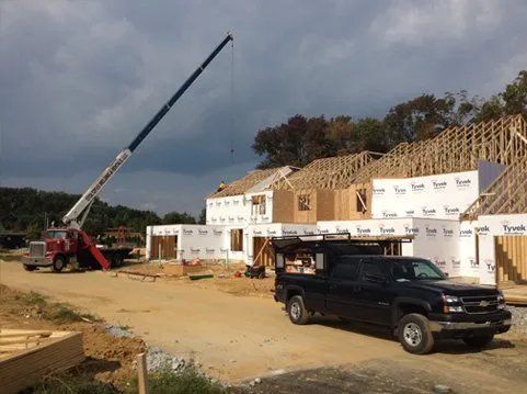 Construction site with a crane lifting materials to frame a house; truck parked in foreground.