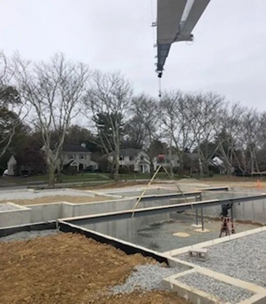 Construction site with foundation, gravel, crane overhead, and houses in the background. Overcast sky.