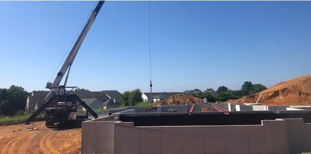 Crane lifting a large black object over a concrete foundation on a construction site. Clear blue sky.