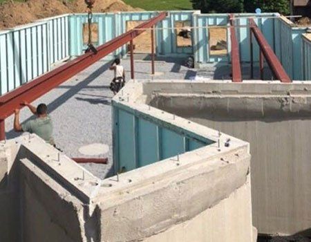 Construction site with workers positioning red metal beams over a concrete foundation with blue insulated walls.