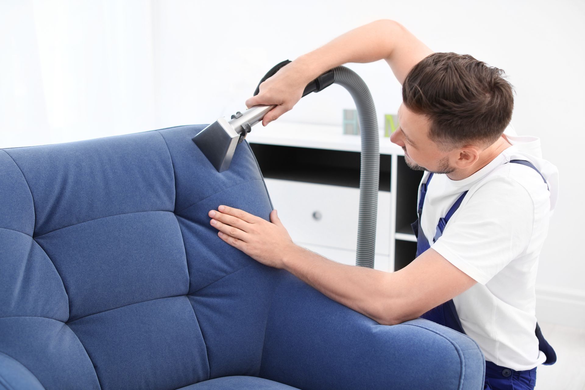 A man is cleaning a blue couch with a vacuum cleaner.