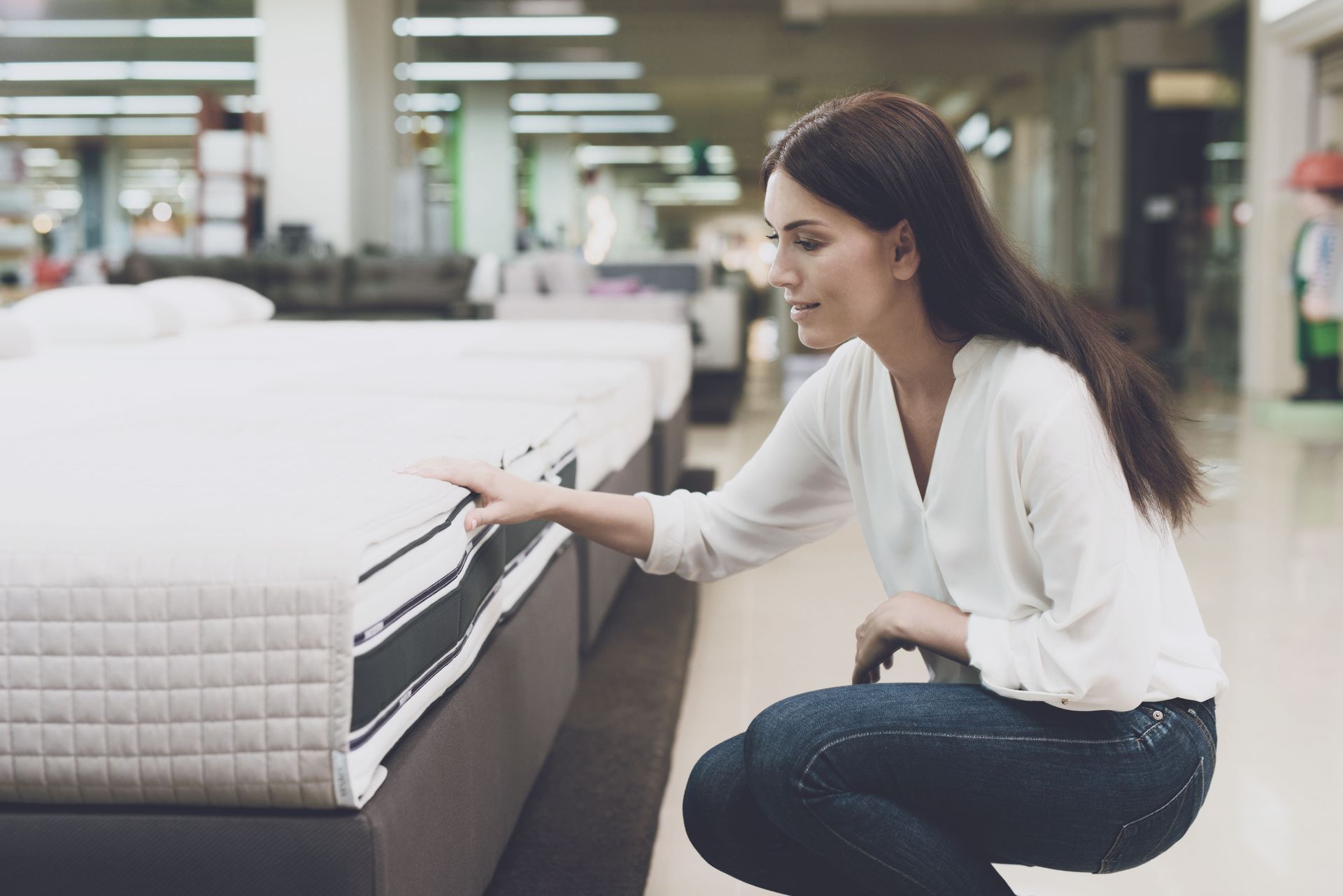 A woman is kneeling down in front of a mattress in a store.