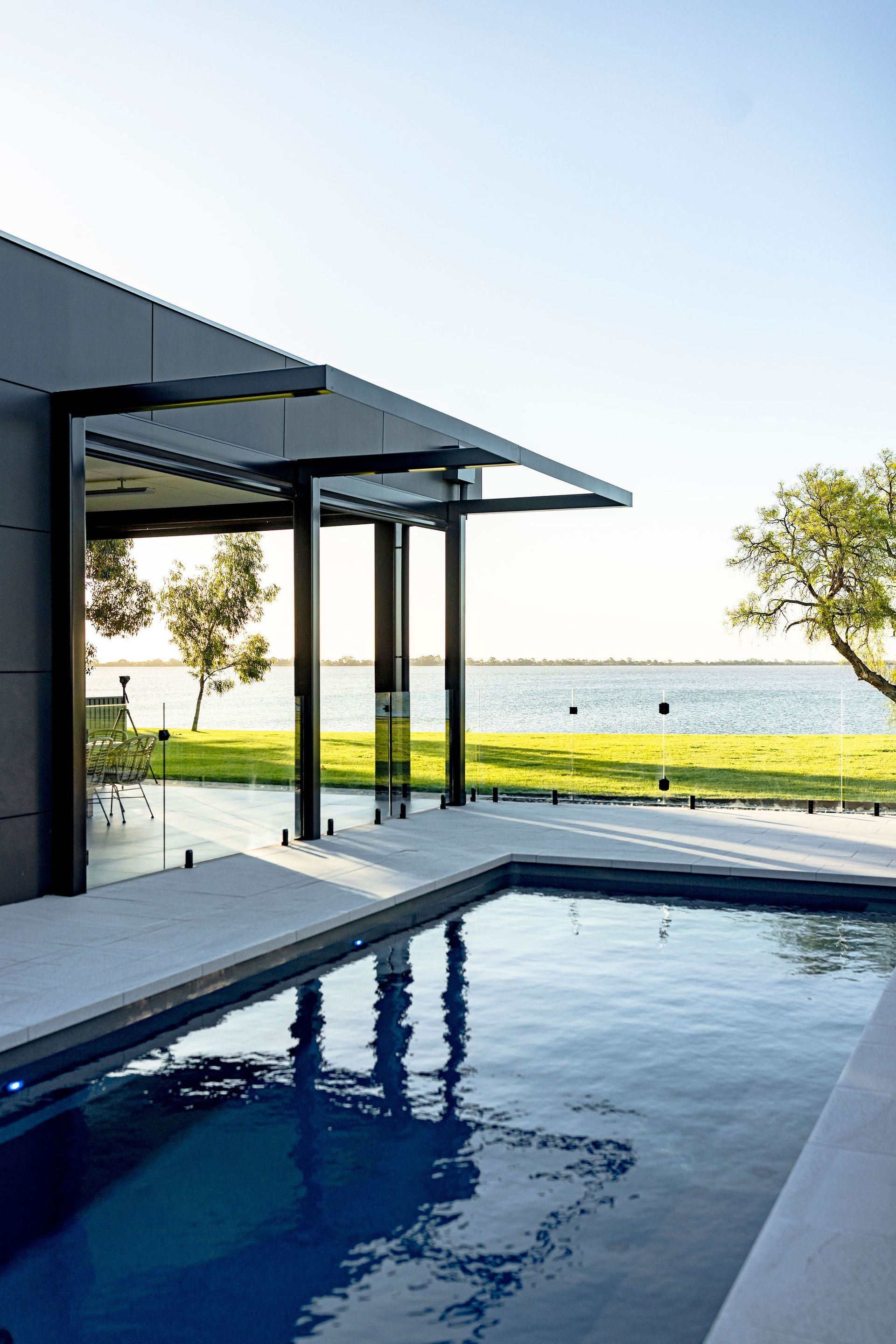 Modern home with pool overlooking a body of water. Black canopy, blue pool, green grass, and trees — Swan Hill Pool & Spa Centre in Swan Hill, VIC