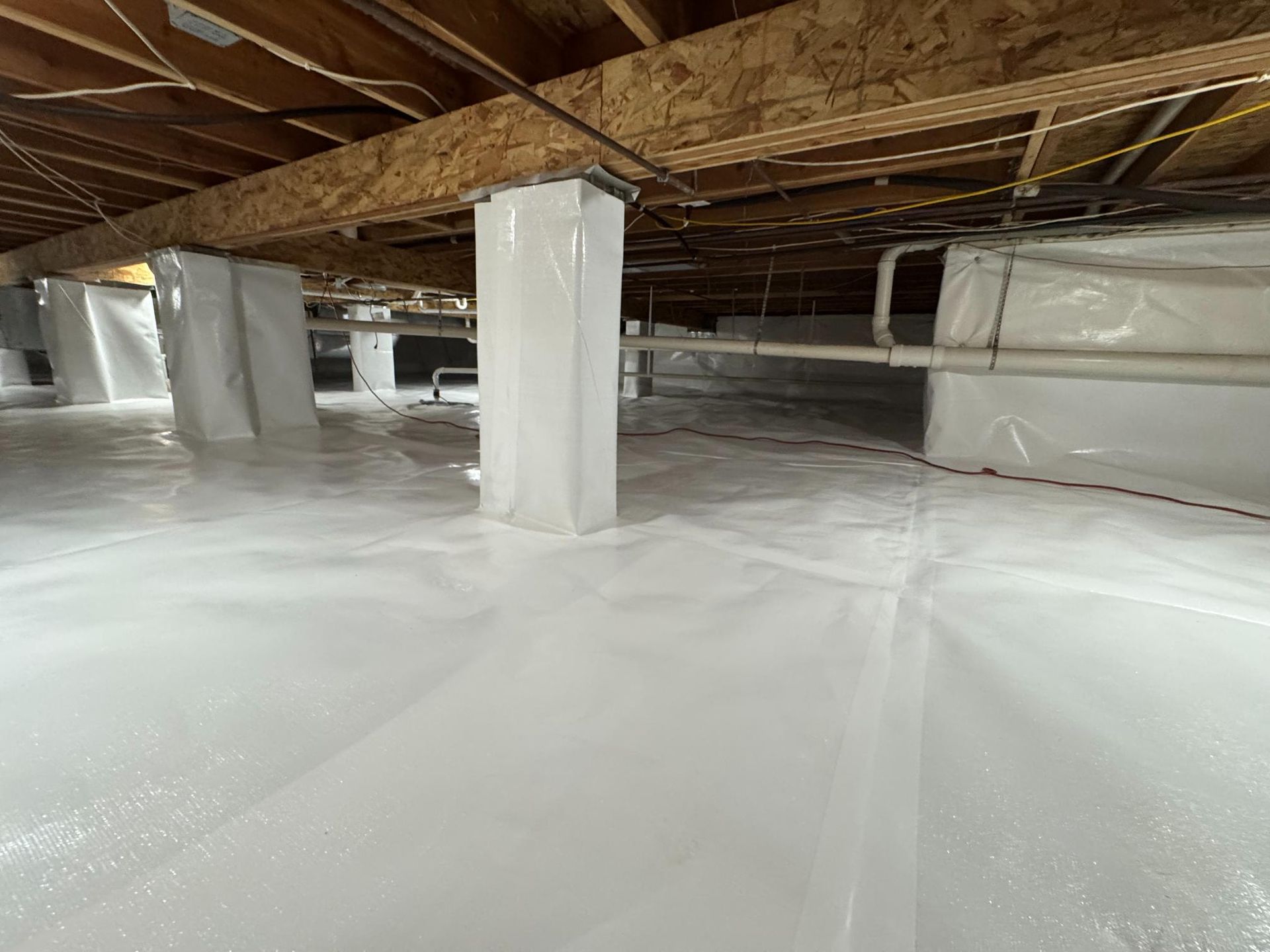 A crawl space encapsulated with white vapor barrier plastic wrapping the floor and support pillars under floor joists.