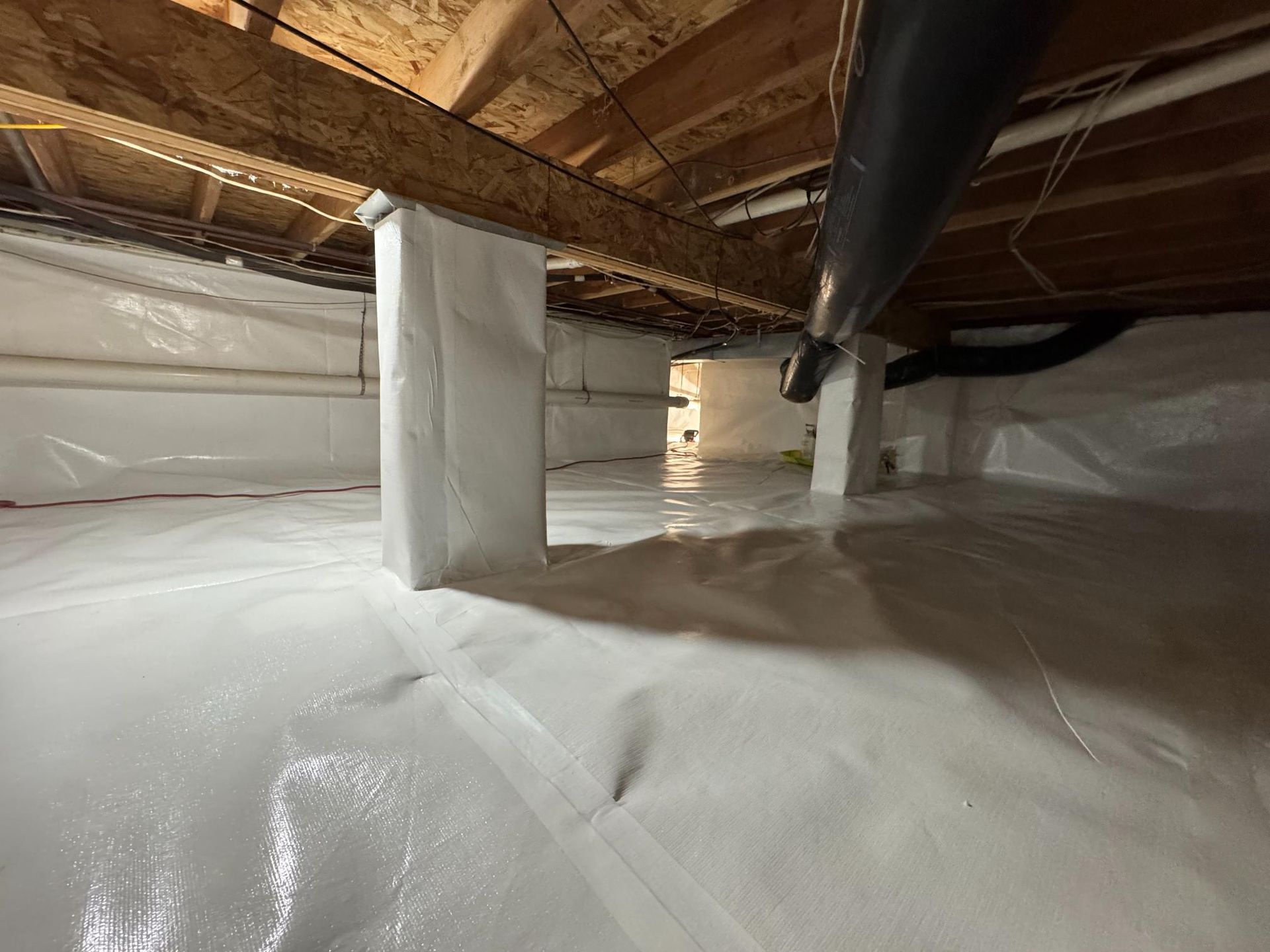A crawl space with white vapor barrier lining the floor and wrapping around support columns under wooden floor joists.