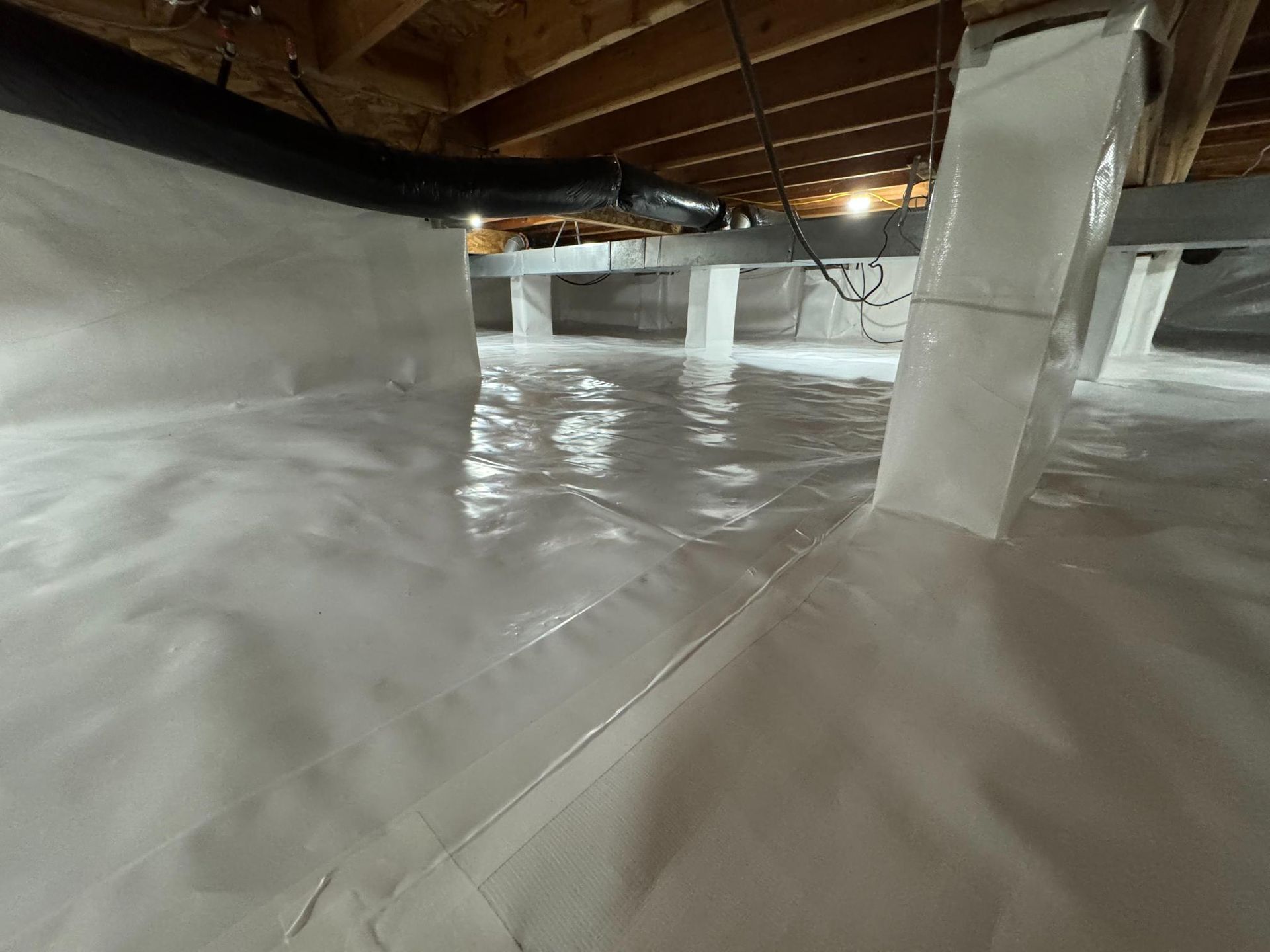 A crawl space featuring a white vapor barrier covering the floor and foundation piers under wooden floor joists.