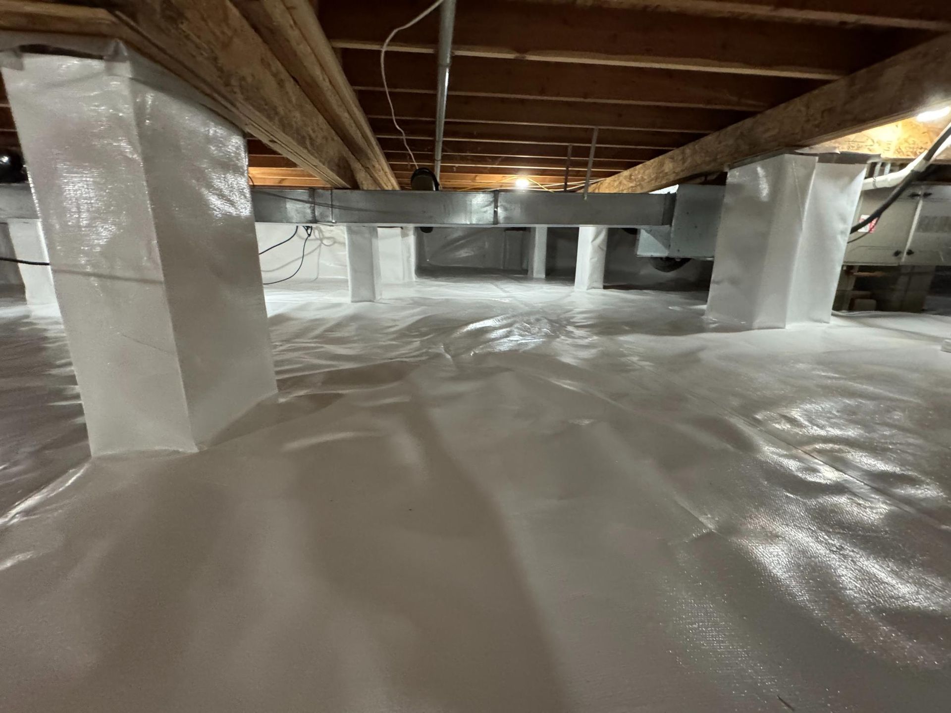 Crawl space with white vapor barrier flooring and wrapped support posts, featuring wooden beams and ductwork overhead.