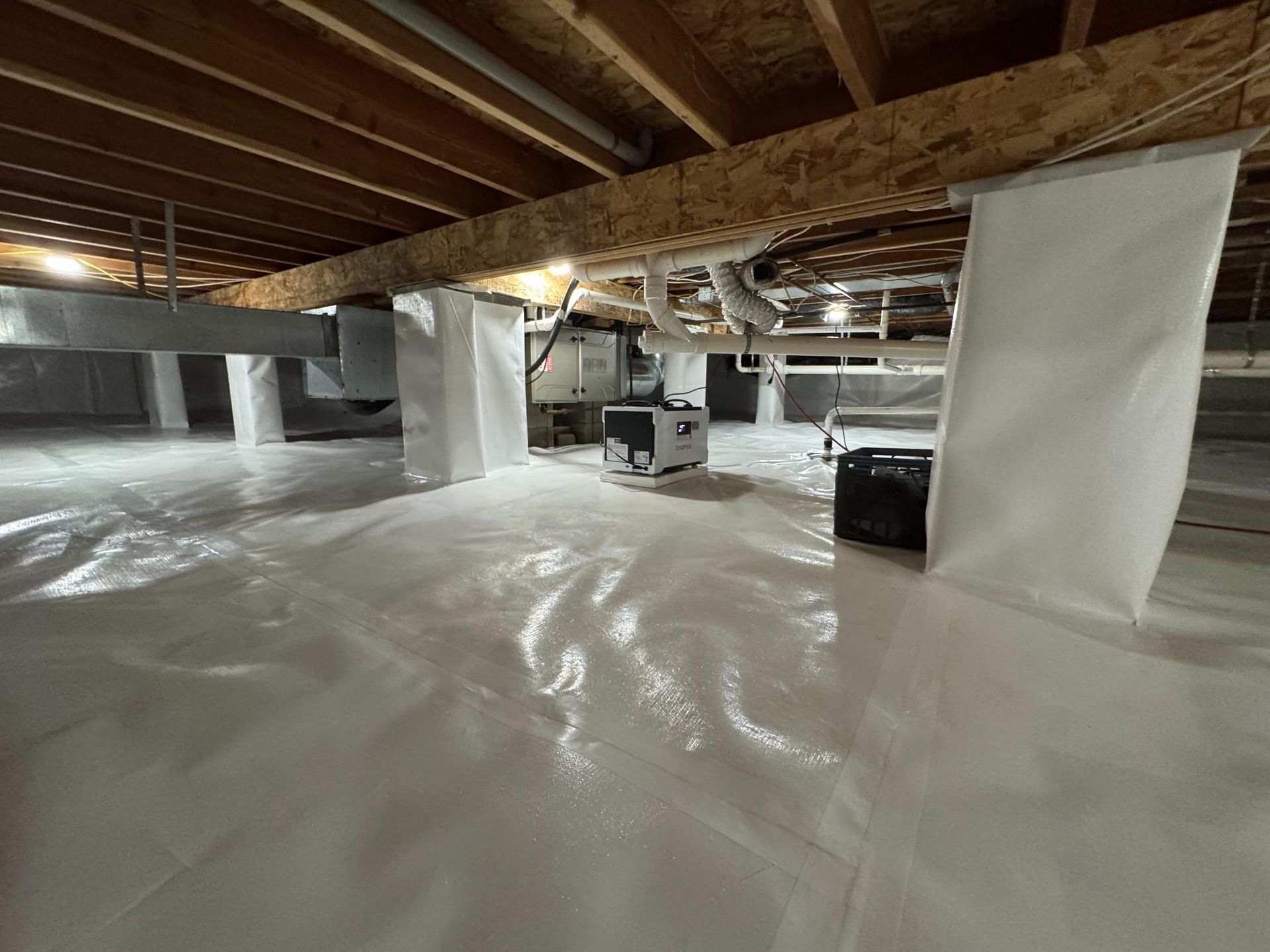 A crawl space with white vapor barrier flooring and pillar wraps, a dehumidifier, and exposed wooden ceiling joists.
