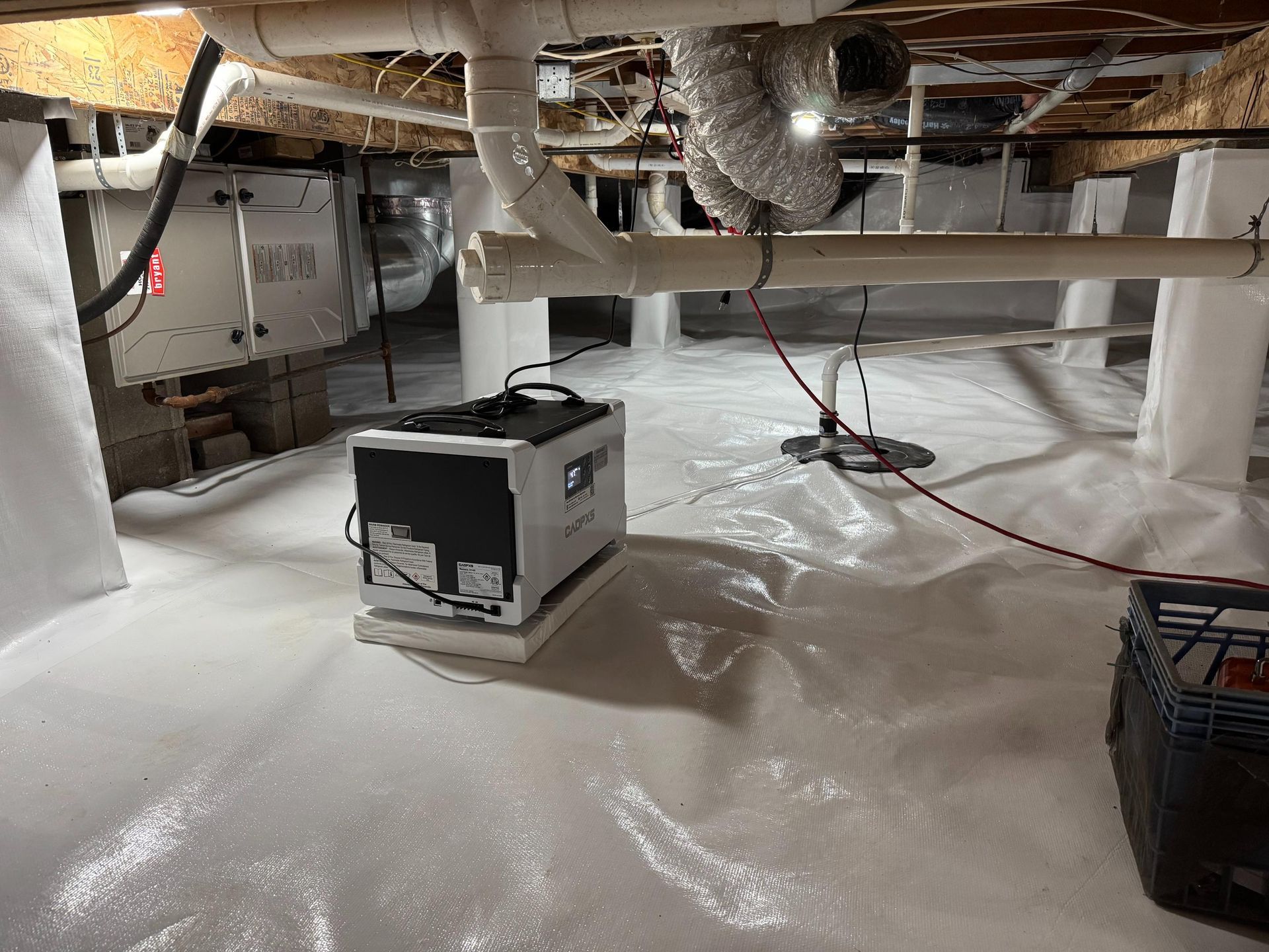 A crawl space encapsulated with white vapor barriers, featuring a central dehumidifier, pipes, and a black crate.