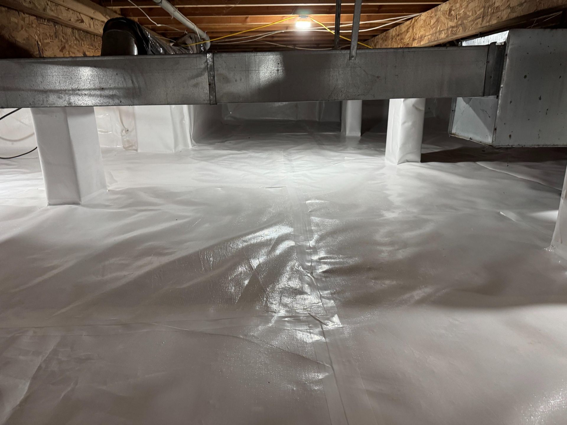 A clean, white vapor barrier covers the floor of a crawl space beneath metallic ductwork and support pillars.