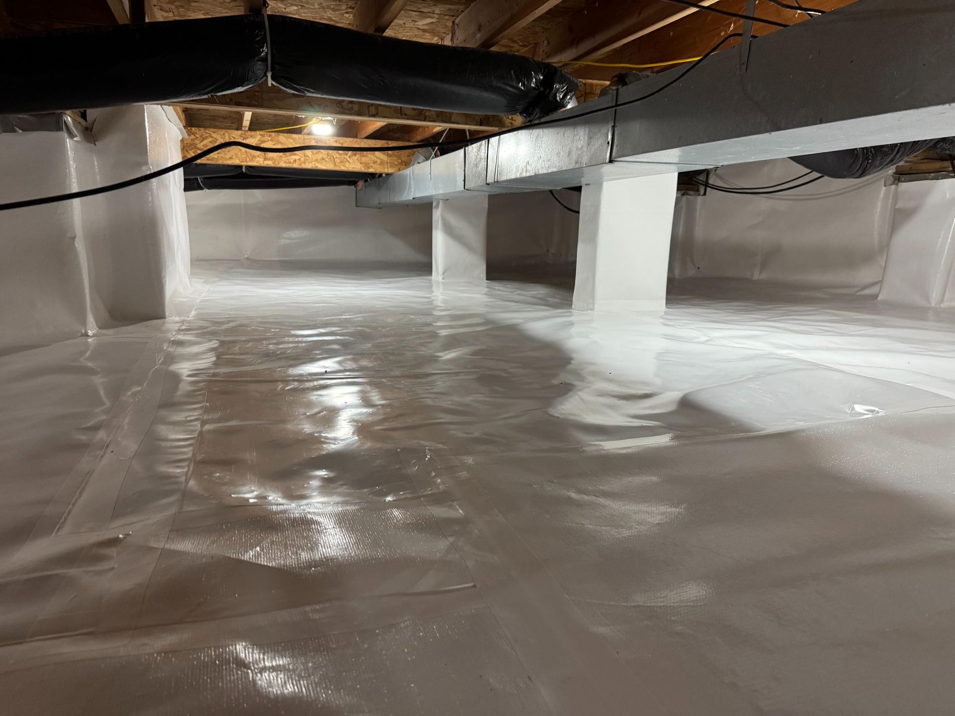 A clean, bright crawl space with white vapor barrier plastic on the ground and walls, featuring insulated metal ductwork.