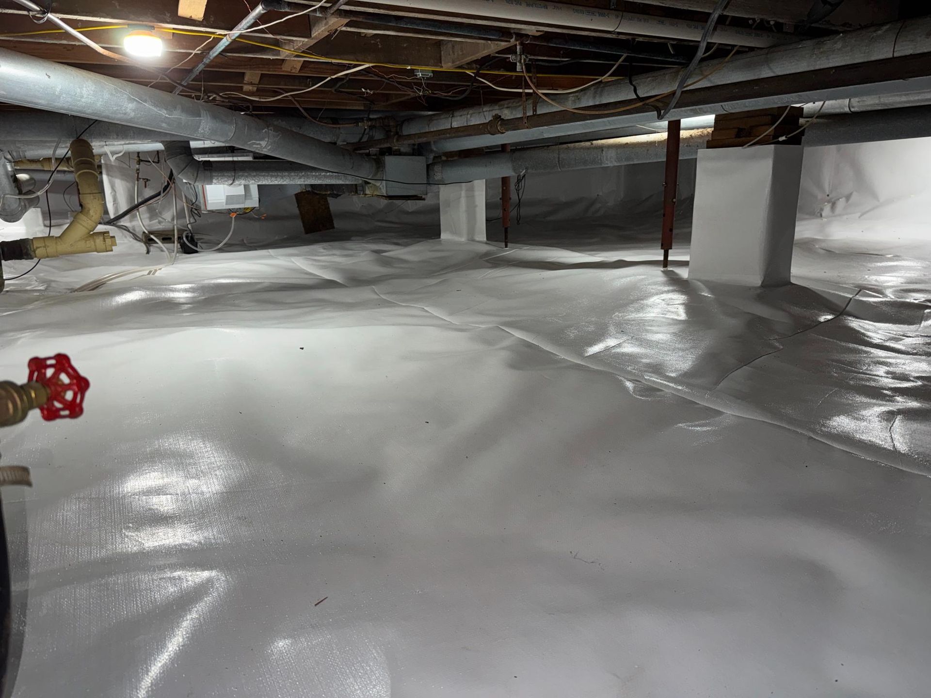 A crawl space with a clean white vapor barrier covering the floor, wrapping around support pillars, and overhead piping.