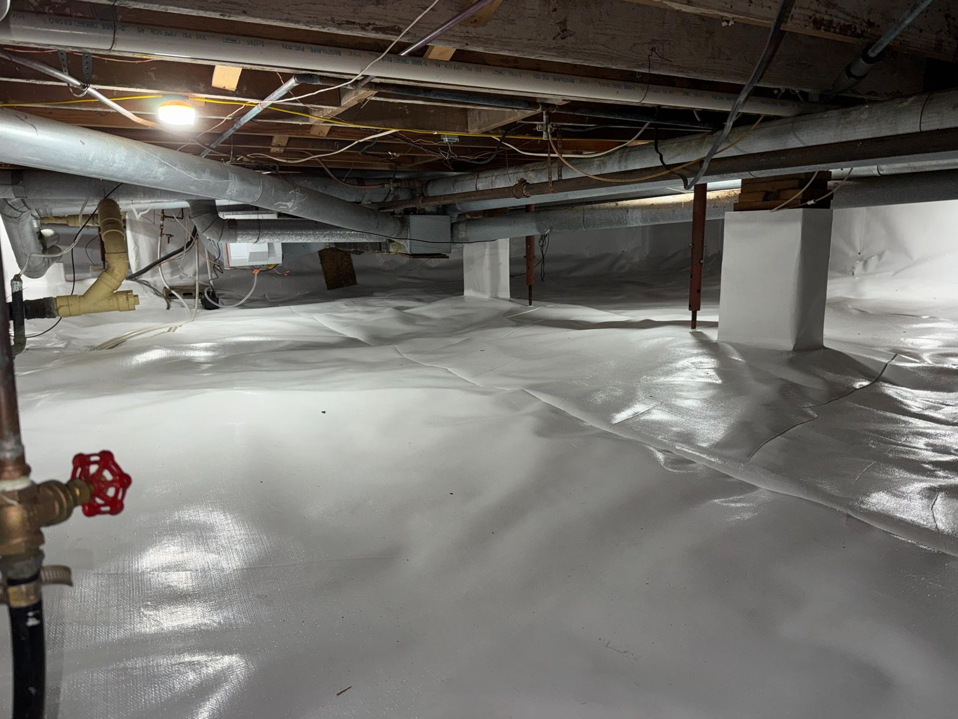 A crawl space with a white vapor barrier covering the ground, white-painted support pillars, and overhead ductwork.