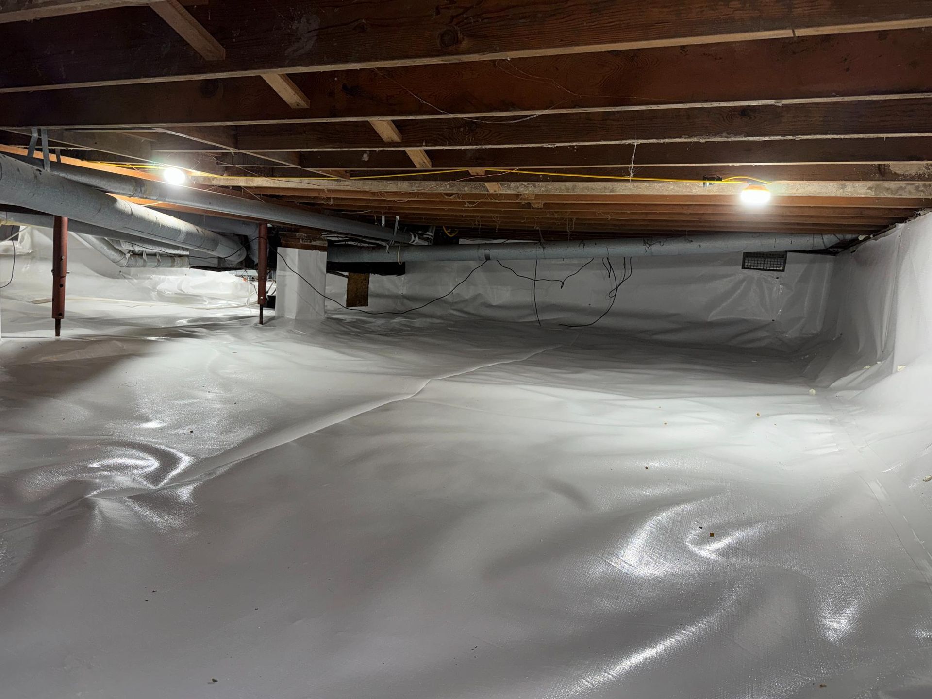 A brightly lit crawl space with a white vapor barrier covering the ground and walls, featuring wooden floor joists above.