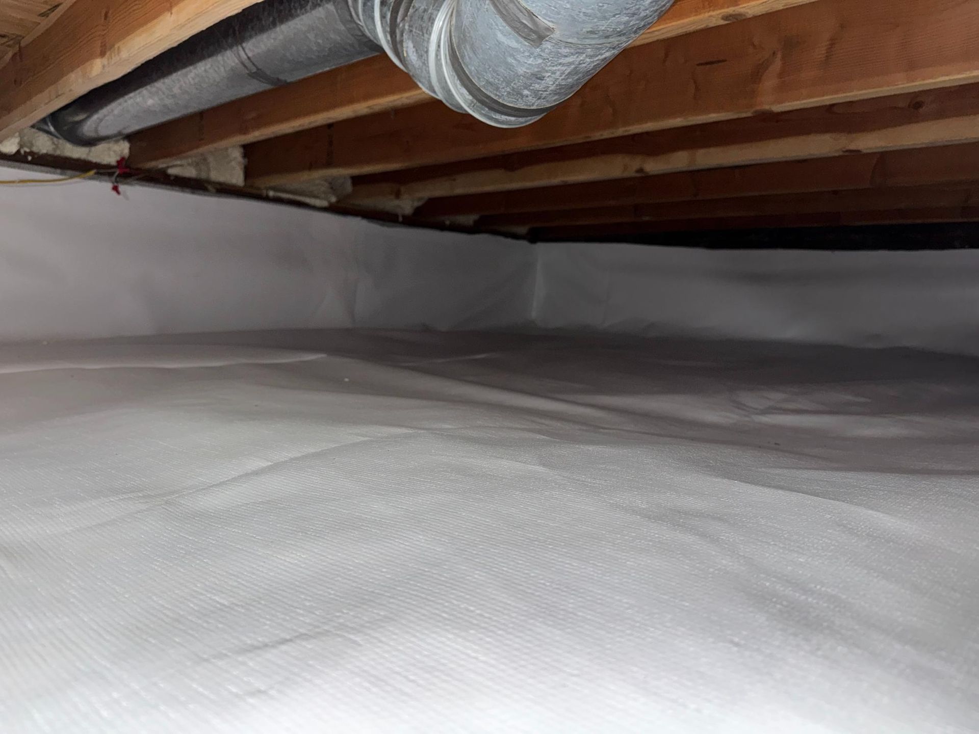 A clean, white vapor barrier lines the ground and walls of a crawl space with wooden floor joists and ductwork above.