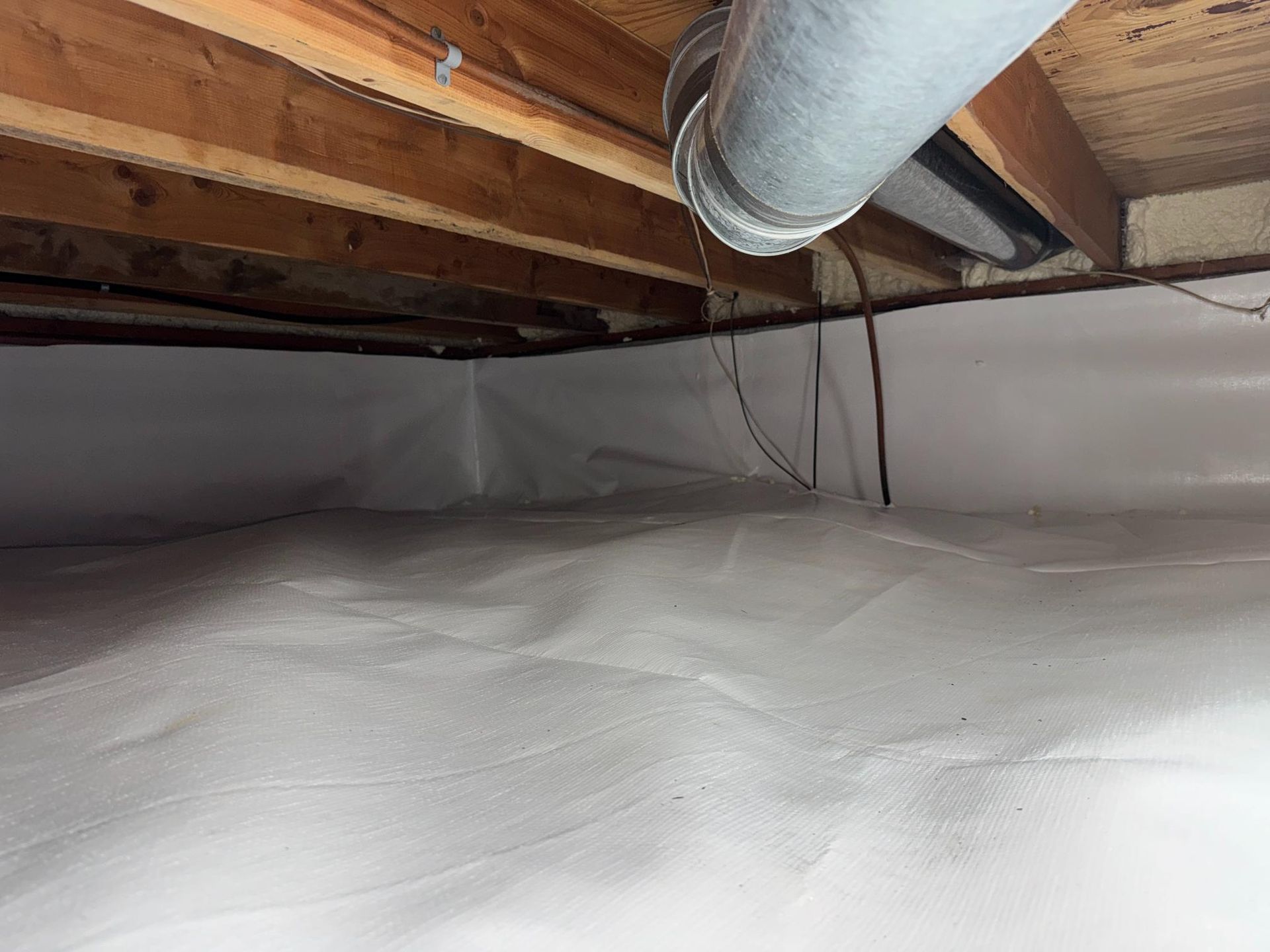 A white vapor barrier installed in a crawl space under wooden floor joists with a metal HVAC duct visible.