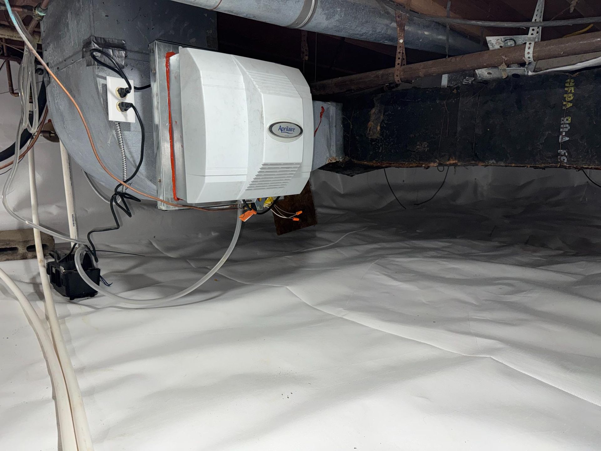 A white Aprilaire humidifier mounted on an HVAC duct in a crawl space with a white vapor barrier on the ground.