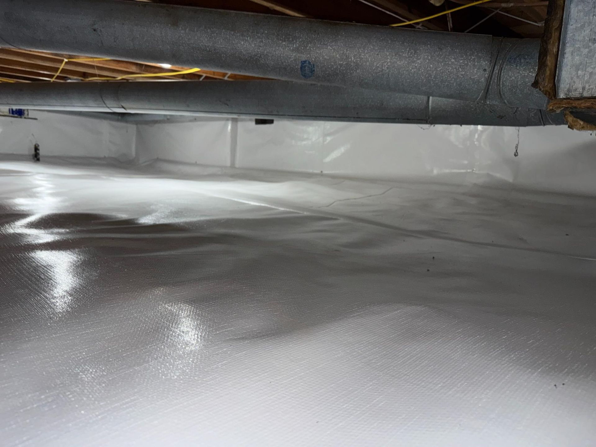 A crawl space with white vapor barrier liner covering the floor and walls and insulated ductwork hanging above.