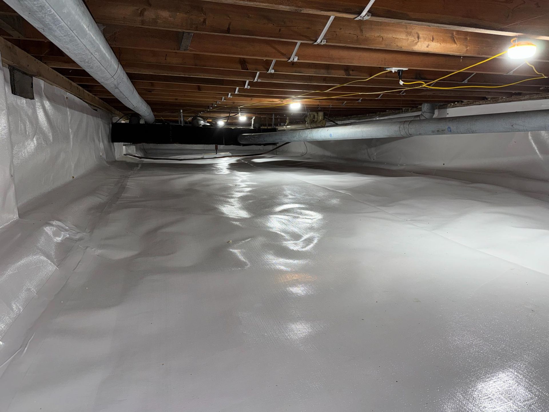 A bright, clean crawlspace featuring a white vapor barrier covering the floor and walls under wooden ceiling joists.