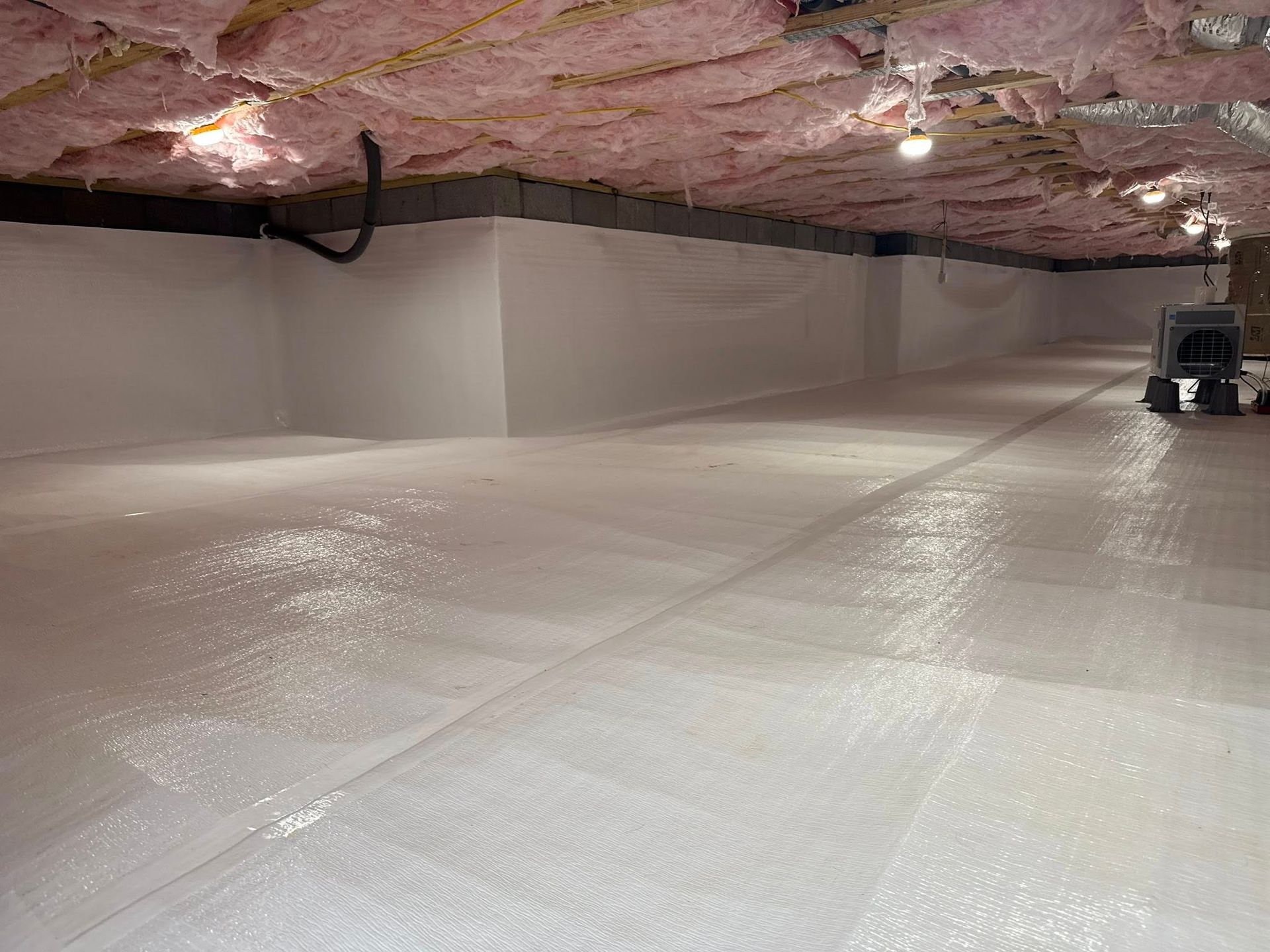 A crawl space with a clean white vapor barrier on the floor, white walls, and pink insulation between the ceiling joists.