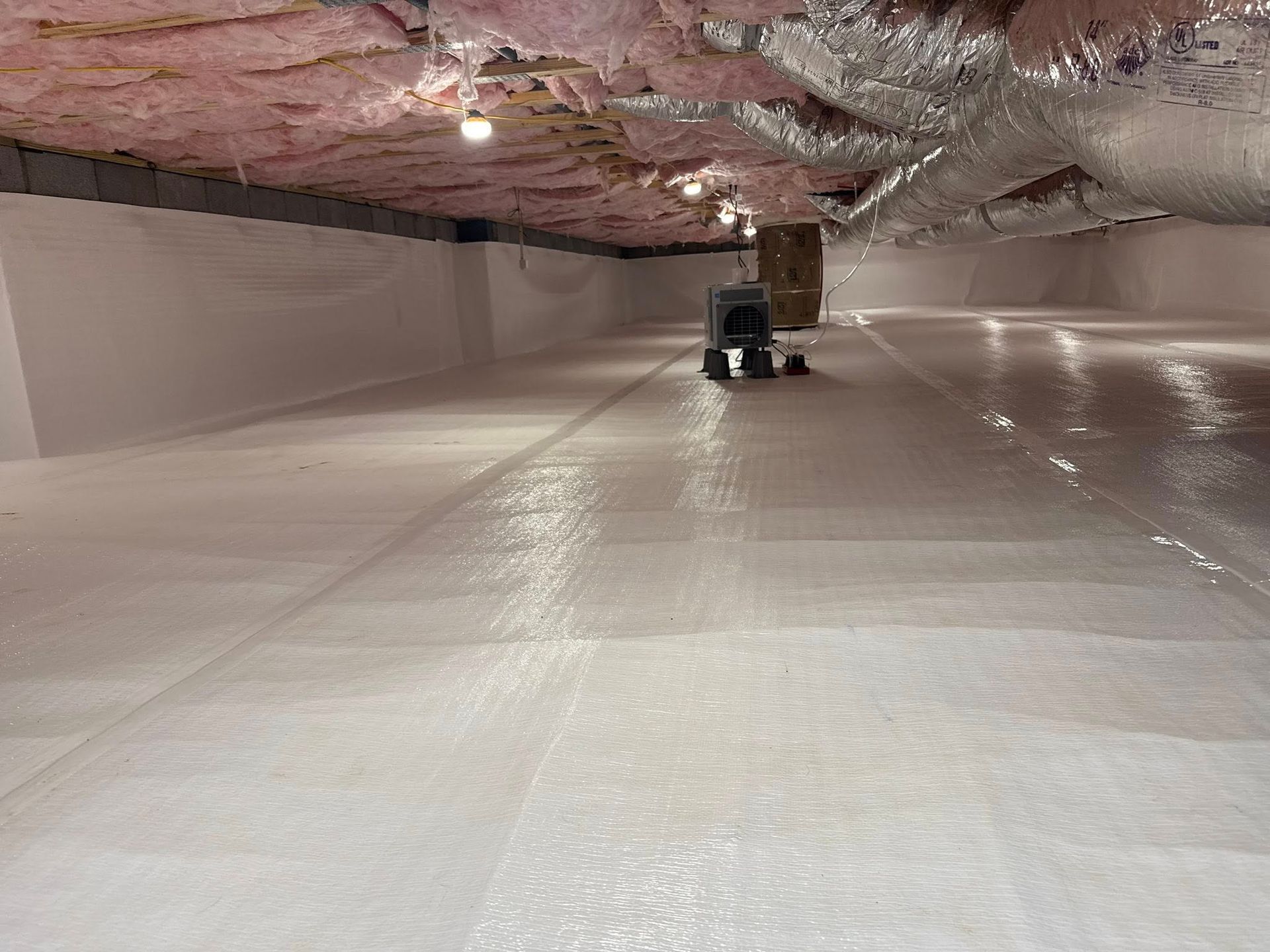 A crawl space with white vapor barrier flooring, pink insulation on the ceiling, and a dehumidifier in the center.